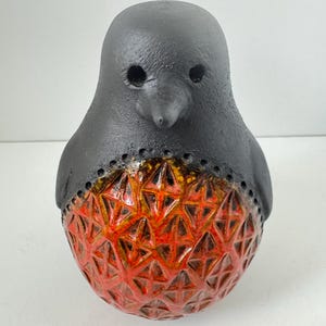 Può includere: Una statuetta di uccello in ceramica con testa e parte superiore del corpo nero opaco caratterizzate da due rientranze oculari circolari. La parte inferiore arrotondata è rifinita con uno smalto arancione e rosso vivace con una trama a rombi.