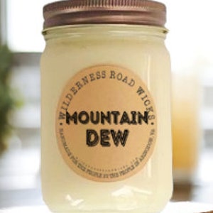 May include: A clear glass jar candle with a brown label that reads "Wilderness Road Wicks Mountain Dew" in black text. The candle is on a wooden surface.