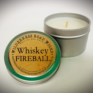 May include: A silver tin candle with a green and brown label that reads "Wilderness Road Wicks Natural Soy Candle Whiskey Fireball Handmade for the People by the People in Abingdon, VA wildernessroadwicks.com".