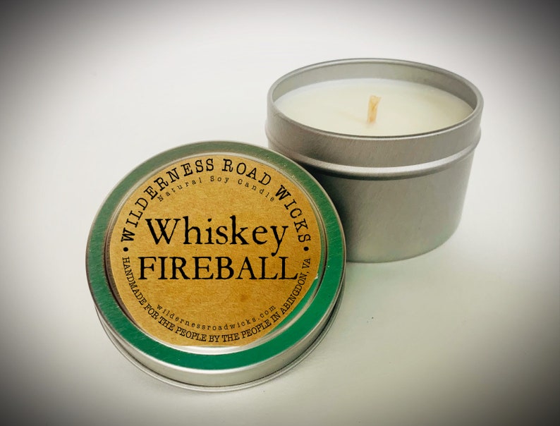 May include: A silver tin candle with a green and brown label that reads "Wilderness Road Wicks Natural Soy Candle Whiskey Fireball Handmade for the People by the People in Abingdon, VA wildernessroadwicks.com".