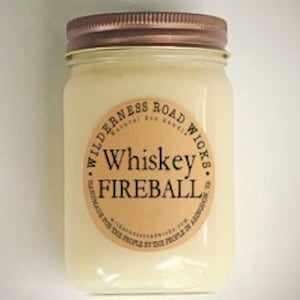 May include: A clear glass jar candle with a copper lid. The label reads "Wilderness Road Wicks Natural Soy Goods Whiskey Fireball Handmade for the People by the People in America".