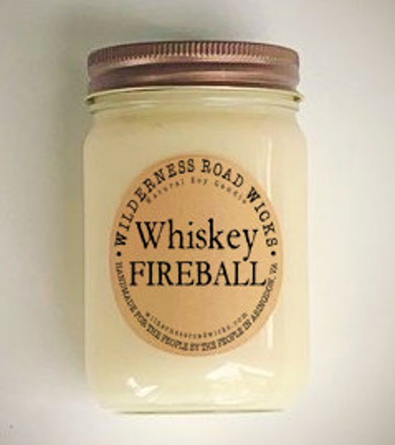 May include: A clear glass jar candle with a copper lid. The label reads "Wilderness Road Wicks Natural Soy Goods Whiskey Fireball Handmade for the People by the People in America".