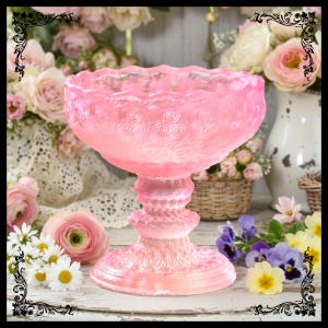 May include: A pink, ornate glass pedestal bowl. The bowl has a scalloped rim and a detailed, swirling pattern. The base is also intricately designed. The bowl is surrounded by flowers, including roses and daisies.