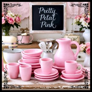 May include: A collection of pink ceramic dishware, including cups, saucers, bowls, and a pitcher, arranged on a distressed white surface. A chalkboard sign with the text "Pretty Petal Pink" is in the background, along with floral arrangements and a stand mixer.