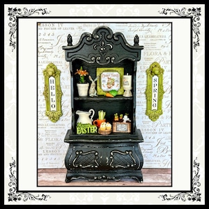 May include: A black painted miniature hutch with two shelves. The top shelf has a floral arrangement, a bunny figurine, and a framed picture that says "Carrot Patch". The bottom shelf has a pitcher, a bowl of carrots, and a sign that says "Happy Easter". The hutch has two green signs on the sides, one that says "Hello" and one that says "Spring".