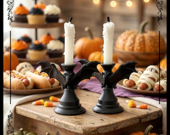 Könnte beinhalten: Zwei schwarze, fledermausförmige Kerzenhalter mit weißen Kerzen auf einem Holztisch. Der Tisch ist mit Halloween-Essen dekoriert, darunter Cupcakes, Hotdogs und Kürbisse. Maisbonbons sind auf dem Tisch verstreut.