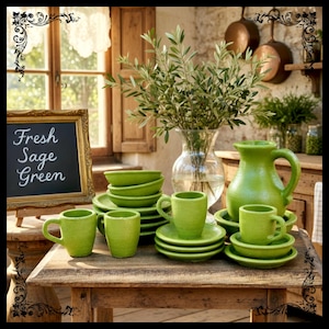 May include: A rustic wooden table displays a collection of sage green ceramic tableware. The set includes cups, saucers, bowls, and a pitcher. A chalkboard sign reads "Fresh Sage Green". A vase of olive branches adds a natural touch.