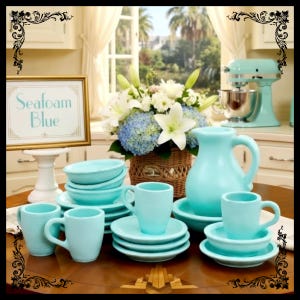 May include: A collection of seafoam blue ceramic tableware, including cups, saucers, bowls, and a pitcher, arranged on a wooden table. A sign reads "Seafoam Blue". A bouquet of white lilies and blue hydrangeas sits in a wicker basket.