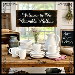 May include: A rustic wooden table displays white dishware, including mugs, plates, and a pitcher. A sign reading "Welcome to The Bramble Hollow" hangs above. A chalkboard reads "Pure White Cotton."