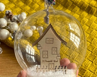 May include: A glass ornament with a wooden house cutout inside. The house has a chimney, windows, and the text "14 Mayfield Street" on the front. The ornament is filled with faux snow and has a plaid ribbon attached.