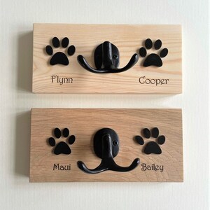 Pode incluir: Dois ganchos de madeira para trela de cachorro com ganchos de metal preto e desenhos de pegadas. O gancho superior é etiquetado como "Flynn" e "Cooper". O gancho inferior é etiquetado como "Maui" e "Bailey".
