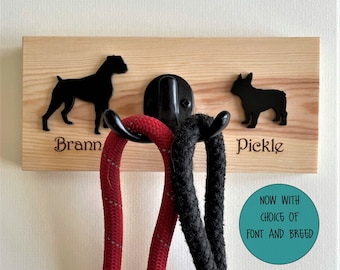 May include: Wooden dog leash rack with two hooks, featuring a black silhouette of a Boxer dog with the name "Brann" and a black silhouette of a French Bulldog with the name "Pickle". Two leashes are hanging on the hooks. The text "NOW WITH CHOICE OF FONT AND BREED" is in a teal circle.