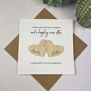 Puede incluir: Una tarjeta blanca con el texto "wishing you both love, laughter and a happily ever after" y dos corazones de madera con los nombres "Jennifer" y "Andrew" grabados en ellos. La tarjeta dice "congratulations on your engagement".