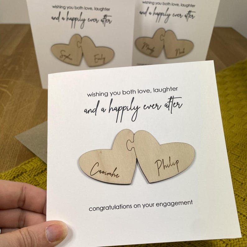 Puede incluir: Una tarjeta blanca con el texto "wishing you both love, laughter and a happily ever after" y dos piezas de rompecabezas de madera en forma de coraz&oacute;n con los nombres "Cavimhe" y "Philip" escritos en ellas. La tarjeta tambi&eacute;n dice "congratulations on your engagement".