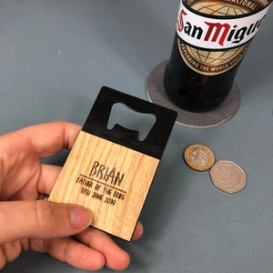Può includere: Apribottiglie in legno con parte superiore in metallo nero. L'apribottiglie è inciso con il testo "BRIAN FATHER OF THE BRIDE 17TH JUNE 2019".
