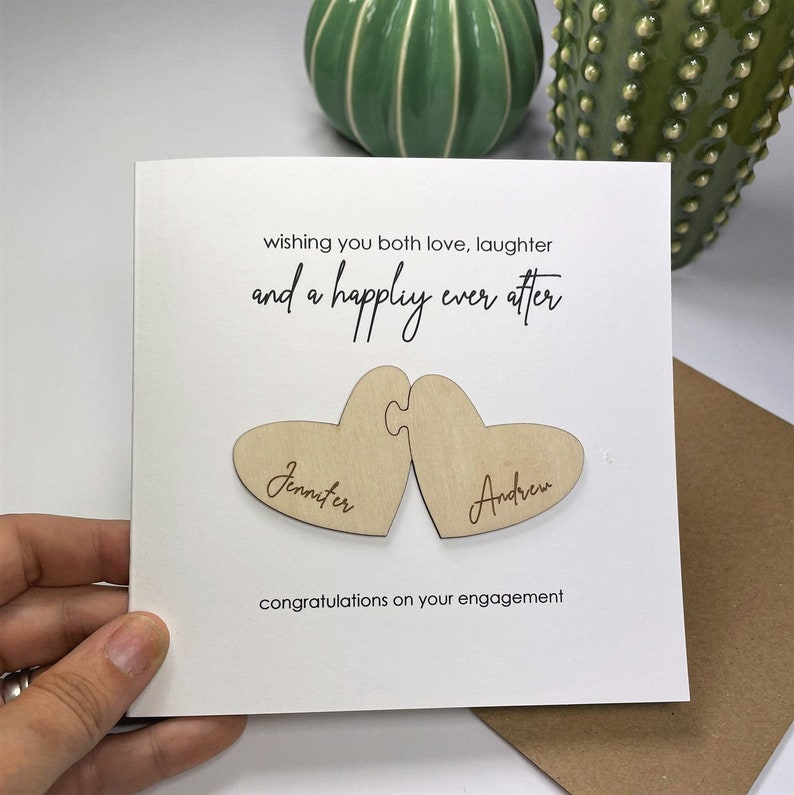 Puede incluir: Una tarjeta blanca con el texto "wishing you both love, laughter and a happily ever after" y dos piezas de rompecabezas de madera en forma de coraz&oacute;n con los nombres "Jennifer" y "Andrew" grabados en ellas. La tarjeta tambi&eacute;n dice "congratulations on your engagement".
