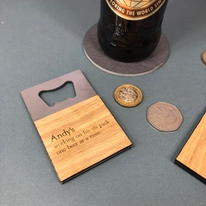 Könnte beinhalten: Ein Flaschenöffner aus Holz und Metall mit dem Text "Andy's working on his six pack one beer at a time." Eine dunkle Bierflasche und Münzen sind im Hintergrund. Der Öffner hat einen silbernen Metallaufsatz und eine hellbraune Holzbasis.