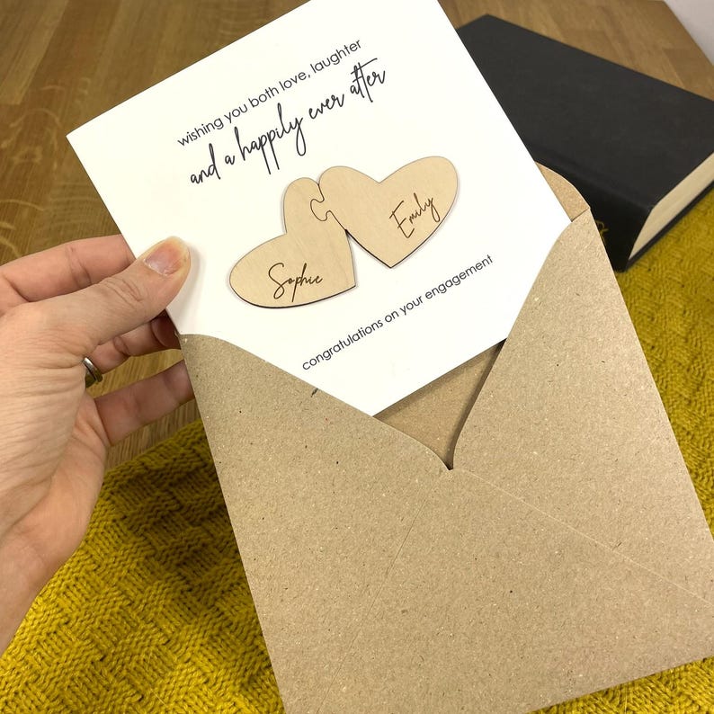 Puede incluir: Una tarjeta blanca con el texto "wishing you both love, laughter and a happily ever after" y dos piezas de rompecabezas en forma de coraz&oacute;n de madera con los nombres "Sophie" y "Emily" inscritos en ellas. La tarjeta est&aacute; en un sobre marr&oacute;n.