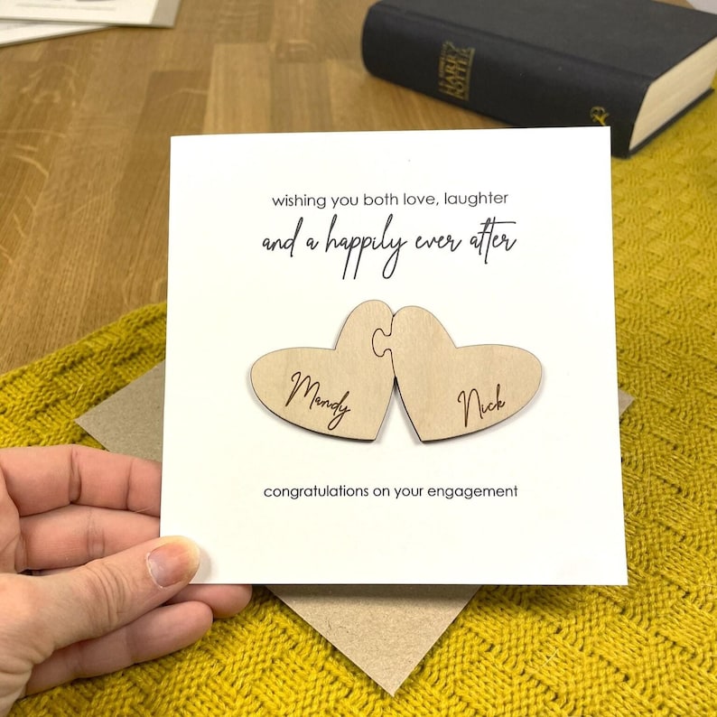 Puede incluir: Una tarjeta blanca con el texto "wishing you both love, laughter and a happily ever after" y dos piezas de rompecabezas de madera en forma de coraz&oacute;n con los nombres "Mandy" y "Nick" en ellas. La tarjeta dice "congratulations on your engagement".