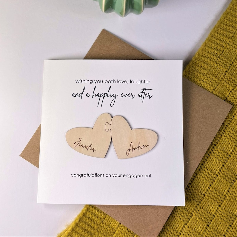 Puede incluir: Una tarjeta blanca con el texto "wishing you both love, laughter and a happily ever after" y dos corazones de madera con los nombres "Jennifer" y "Andrew" grabados en ellos. La tarjeta dice "congratulations on your engagement".