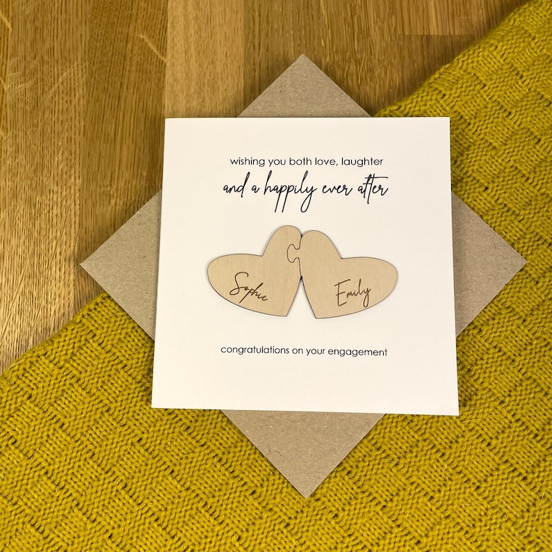 Puede incluir: Una tarjeta blanca con un sobre marr&oacute;n. La tarjeta tiene el texto "wishing you both love, laughter and a happily ever after" con dos corazones de madera que dicen "Sophie" y "Emily". La tarjeta tambi&eacute;n dice "congratulations on your engagement".
