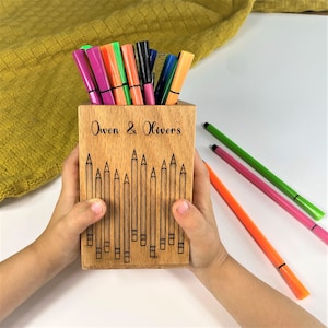 May include: A wooden pencil holder with the text "Owen & Olivers" and pencil illustrations. The holder contains various colored markers. Three additional markers in pink, green, and orange are placed to the side.