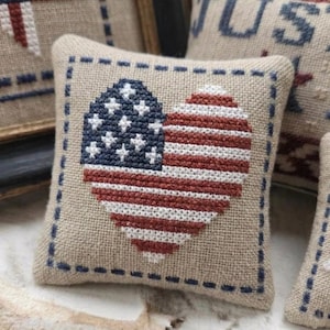 May include: A square beige fabric pillow featuring a cross-stitched heart design in the center. The heart displays the American flag pattern with a blue field of white stars on the left and horizontal red and white stripes on the right side.