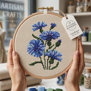 May include: A cross-stitch kit featuring a blue cornflower design. The embroidery is held in a wooden hoop, with the text "Stitched Blossoms" on a tag. The kit includes embroidery floss in shades of blue and green. The text "Artisan Crafts" is visible.
