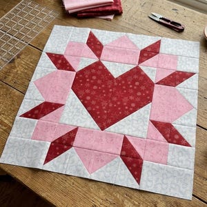 May include: A square quilt block featuring a central dark red heart with a white snowflake pattern, surrounded by light pink and dark red geometric star points on a white background with a subtle gray snowflake print throughout the fabric.