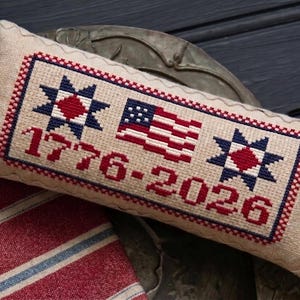 May include: A rectangular, beige cross-stitch pillow with a patriotic design. It features the American flag, two stars, and the numbers "1776-2026" in red and blue thread. The pillow is placed on a decorative tray.