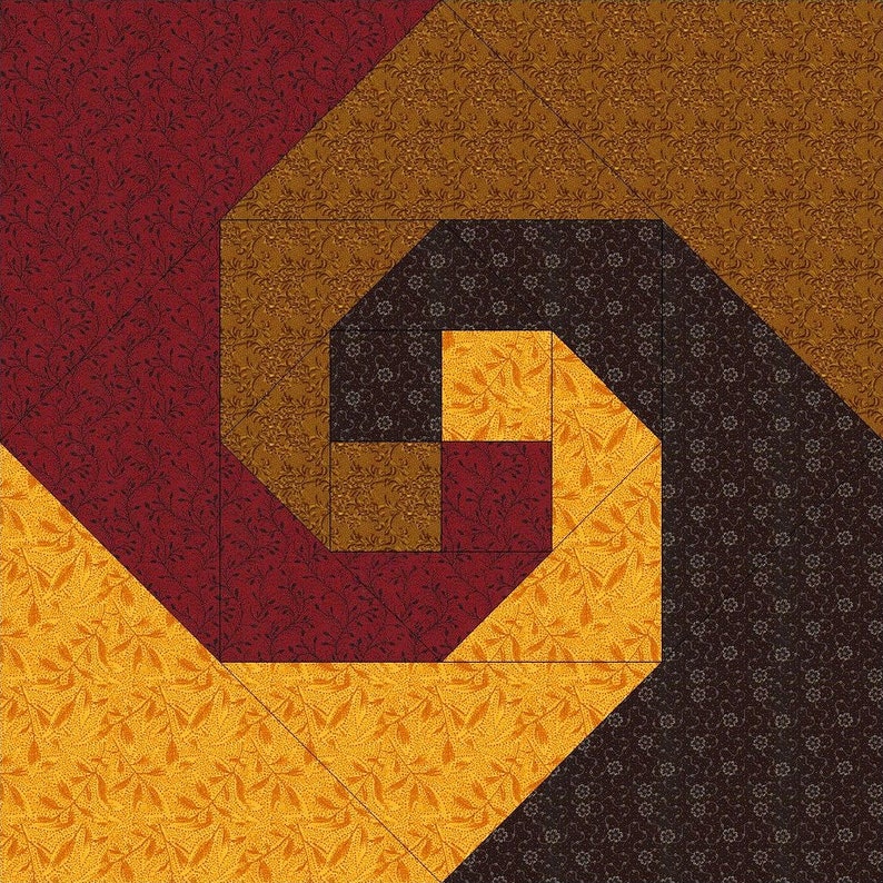 May include: A square quilt block featuring a spiral pattern constructed from fabric pieces in shades of deep red, golden yellow, brown, and dark chocolate. Each section displays a subtle, repeating floral or leaf print across the surface.