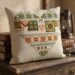 May include: A square, beige pillow with a heart-shaped cross-stitch design. The design features geometric patterns in shades of green, orange, and brown. A small, hanging heart is also part of the design. The pillow is placed on a wooden surface.