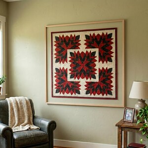 May include: A framed quilt with a repeating floral pattern in red and black on a cream background. The quilt is displayed on a light green wall. The frame is a light wood color.