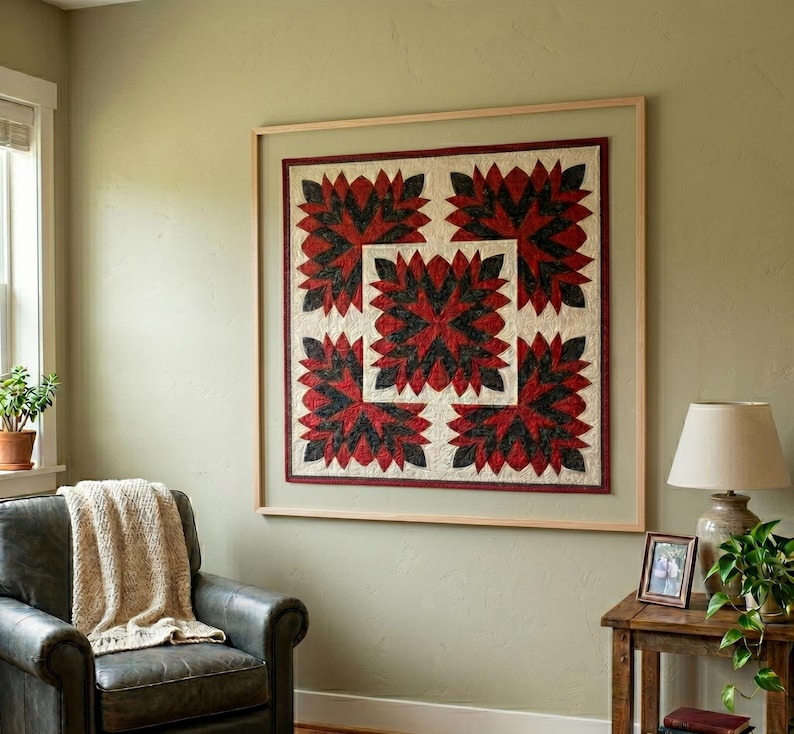 May include: A framed quilt with a repeating floral pattern in red and black on a cream background. The quilt is displayed on a light green wall. The frame is a light wood color.