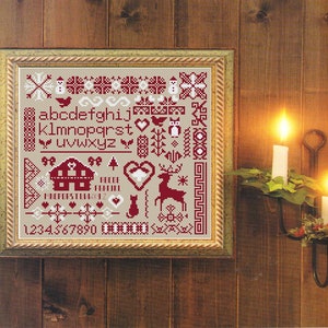 May include: A framed cross-stitch sampler with a beige background and red stitching. The sampler features a variety of Christmas-themed designs, including a house, a reindeer, a heart, a cat, a snowman, a bird, a tree, and the alphabet. The numbers 1234567890 are also included in the design.