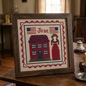 May include: Framed cross-stitch artwork with a red house, a woman in a red dress, and the word "June" with American flags. The artwork is in a dark brown wooden frame, suitable for home decoration.