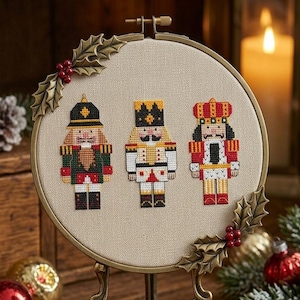 May include: A cross-stitch embroidery hoop featuring three toy soldier nutcrackers. The nutcrackers are in red, green, and gold uniforms. The hoop is decorated with holly leaves and berries, and is displayed on a wooden surface.