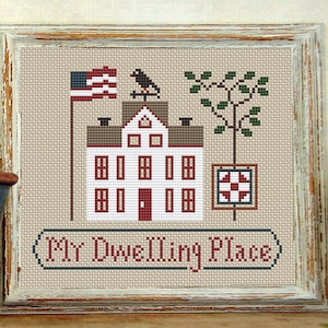 Puede incluir: Obra de arte de punto de cruz de una casa blanca con adornos rojos, una bandera estadounidense y una veleta de cuervo. También se muestra un árbol y un bloque de colcha. El texto "My Dwelling Place" está bordado debajo de la casa.