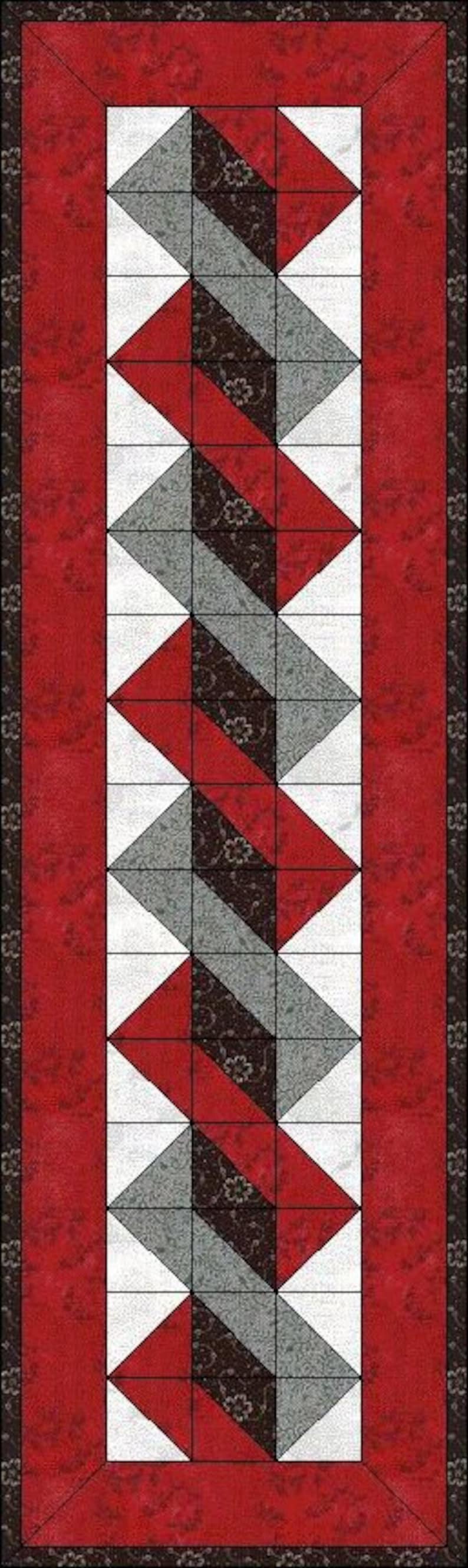 May include: A rectangular table runner featuring a central column of geometric quilt blocks in red, grey, and black floral patterns, bordered by white triangles. The entire design is framed by a wide red border with a dark floral print.