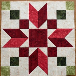 May include: A square quilt block featuring a central dark red square surrounded by a star pattern made of deep red and lighter red diamond-shaped fabric pieces. The background is cream with a subtle leaf print, accented by dark green corner squares.