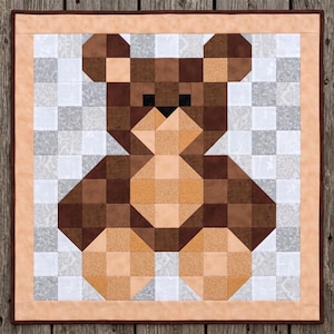 Puede incluir: Una colcha cuadrada con un diseño de oso de peluche pixelado construido a partir de cuadrados de tela en varios tonos de marrón y bronceado. El oso está centrado sobre un fondo de cuadrados estampados en gris claro y blanco, enmarcado por un borde color melocotón.