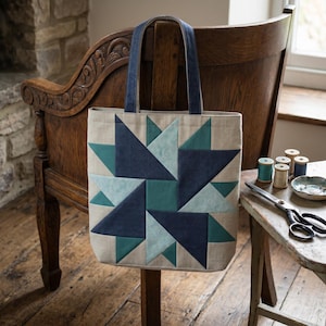 May include: A beige tote bag with a geometric starburst design in shades of teal and navy blue. The bag has navy blue handles and is displayed on a wooden chair. Sewing supplies, including spools of thread and scissors, are on a nearby table.