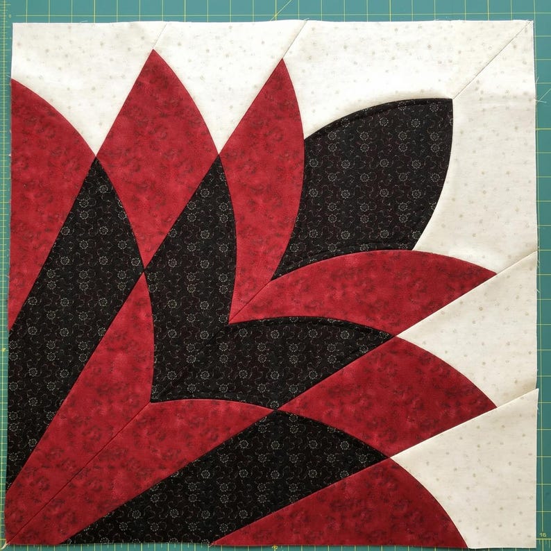 May include: A square quilt block featuring a stylized floral design. The design incorporates red, black, and white fabrics. The red fabric has a solid color, while the black and white fabrics have a subtle pattern. The block is likely part of a larger quilt.