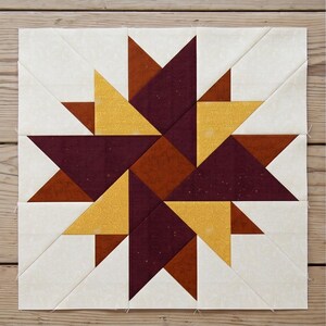 May include: A square quilt block featuring a starburst design. The star is composed of triangles in shades of burgundy, gold, and rust, set against a cream-colored background. The block is made of fabric and is likely part of a larger quilt.