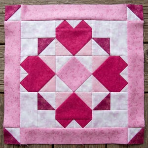 May include: A square quilt block featuring a central design of four dark pink hearts arranged in a cross pattern. The hearts are surrounded by light pink and white geometric fabric pieces, with a solid pink border and dark pink corner squares.
