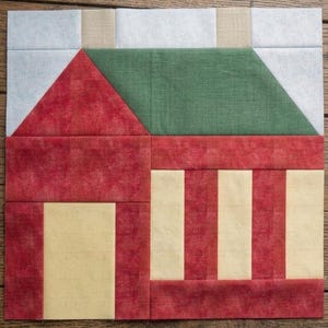 May include: A square quilt block featuring a house design constructed from fabric pieces. The house has a dark red roof, a green upper section, and a lower level with alternating vertical stripes of dark red and light yellow fabric.