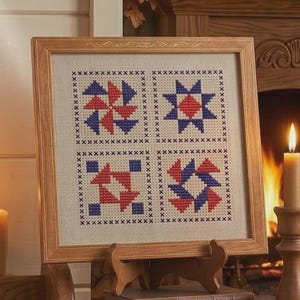 May include: A framed cross-stitch sampler featuring four geometric patterns in red and blue. The sampler is displayed on a wooden easel, with a warm fireplace and candle in the background. The frame is made of light-colored wood.