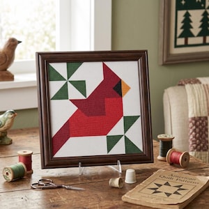 May include: Framed cross-stitch art featuring a red cardinal bird design with green and white geometric accents. The artwork is displayed on a wooden table with sewing supplies and a vintage book titled "American Quilt Patterns".