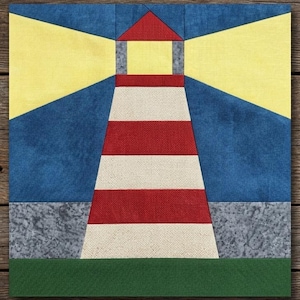Peut inclure: Un bloc de courtepointe en tissu carré représentant un phare central avec des rayures horizontales rouges et crème. Le phare a un toit rouge et un faisceau lumineux jaune s'étendant à gauche et à droite sur un ciel bleu foncé. Une base verte est en dessous.