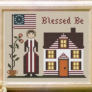 Op de afbeelding: Borduurpatroon met een vrouw in een rood-witte jurk die voor een huis staat met een rood dak en witte muren. Het huis heeft vier ramen en een deur. Op de achtergrond staat een vlaggenmast met een Amerikaanse vlag. De tekst "Blessed Be" is boven het huis geborduurd.